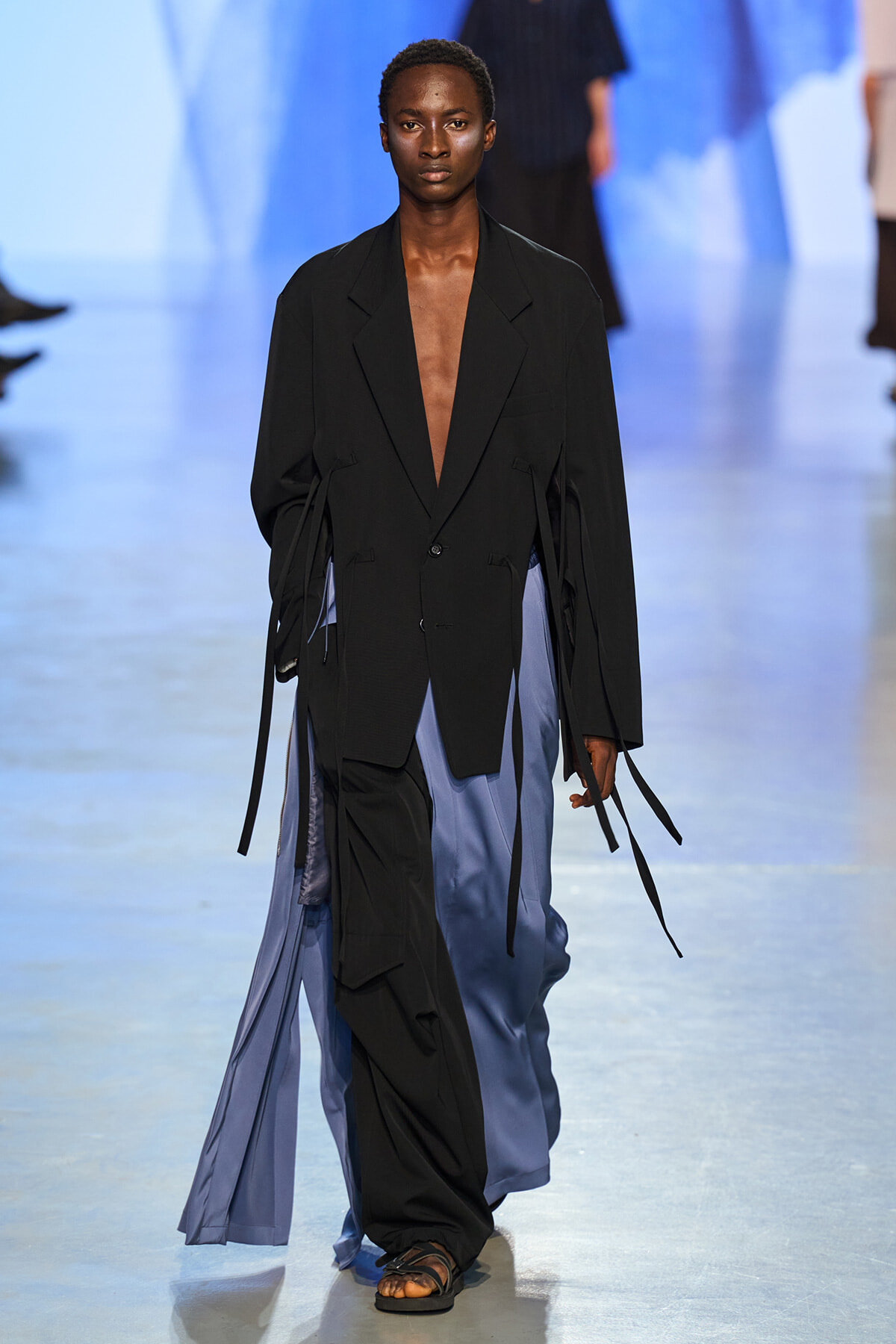 Model walking the runway in a loose black blazer with a deep V, paired with flowing blue satin pants and strappy sandals.