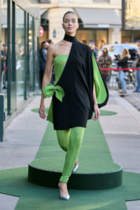 Model walking a street-style runway in a black and lime-green asymmetrical dress, large green bow at the hip, lime tights, and silver heels, outdoor show setting.