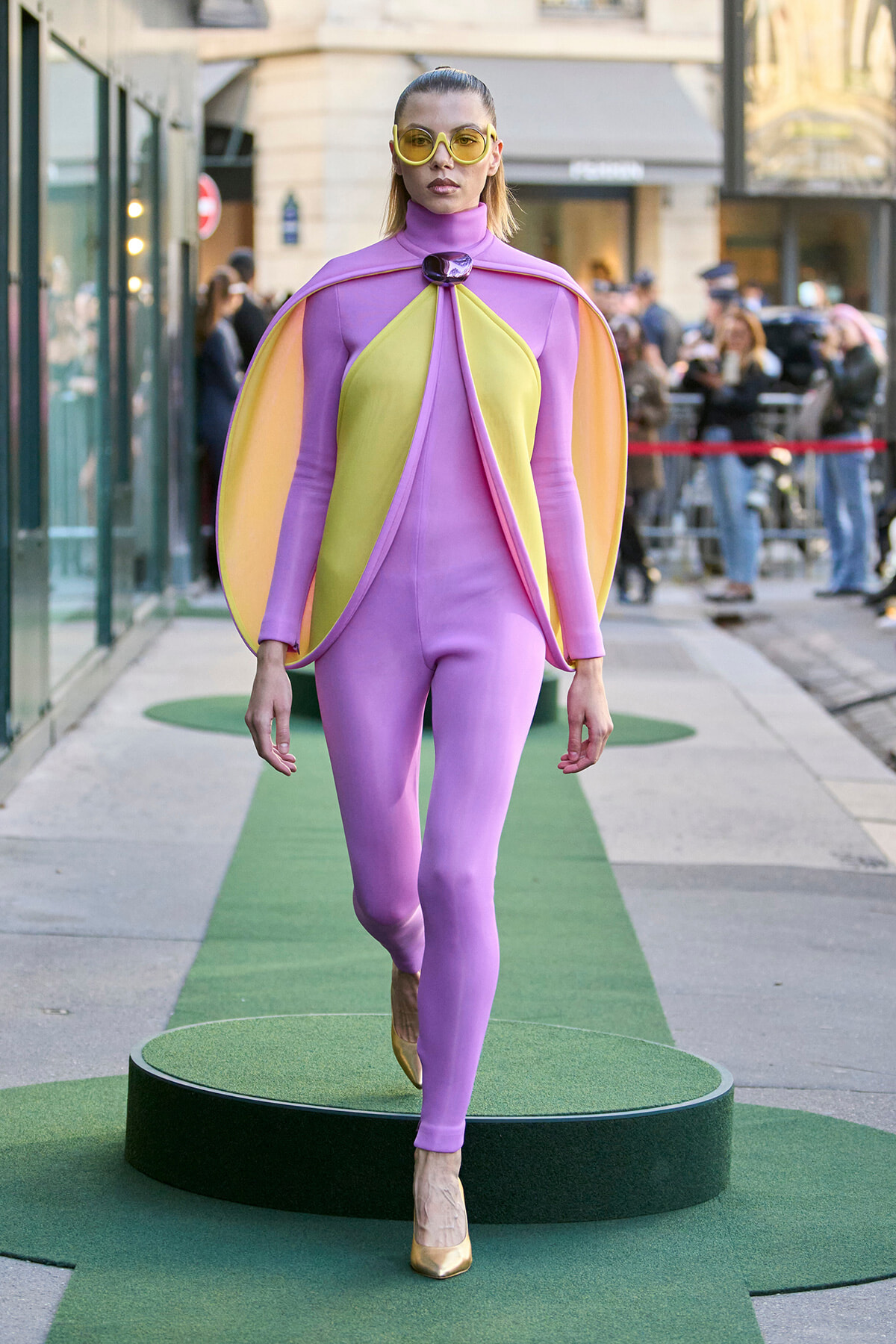 Model wearing a lavender bodysuit with yellow-green cape panels and oversized yellow sunglasses walks a green-carpeted runway.