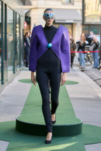 Model in a purple structured blazer over a black bodysuit walks on a green circular runway, wearing oversized reflective sunglasses and a blue pendant.