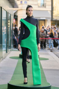 Model wearing a black, long-sleeve bodysuit with a bold green sculptural dress, standing on a green circular runway outdoors and spectators in background.