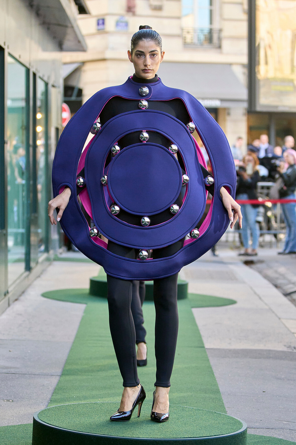 Model walks a street-style runway wearing a bold purple circular dress made of concentric rings and chrome spheres.