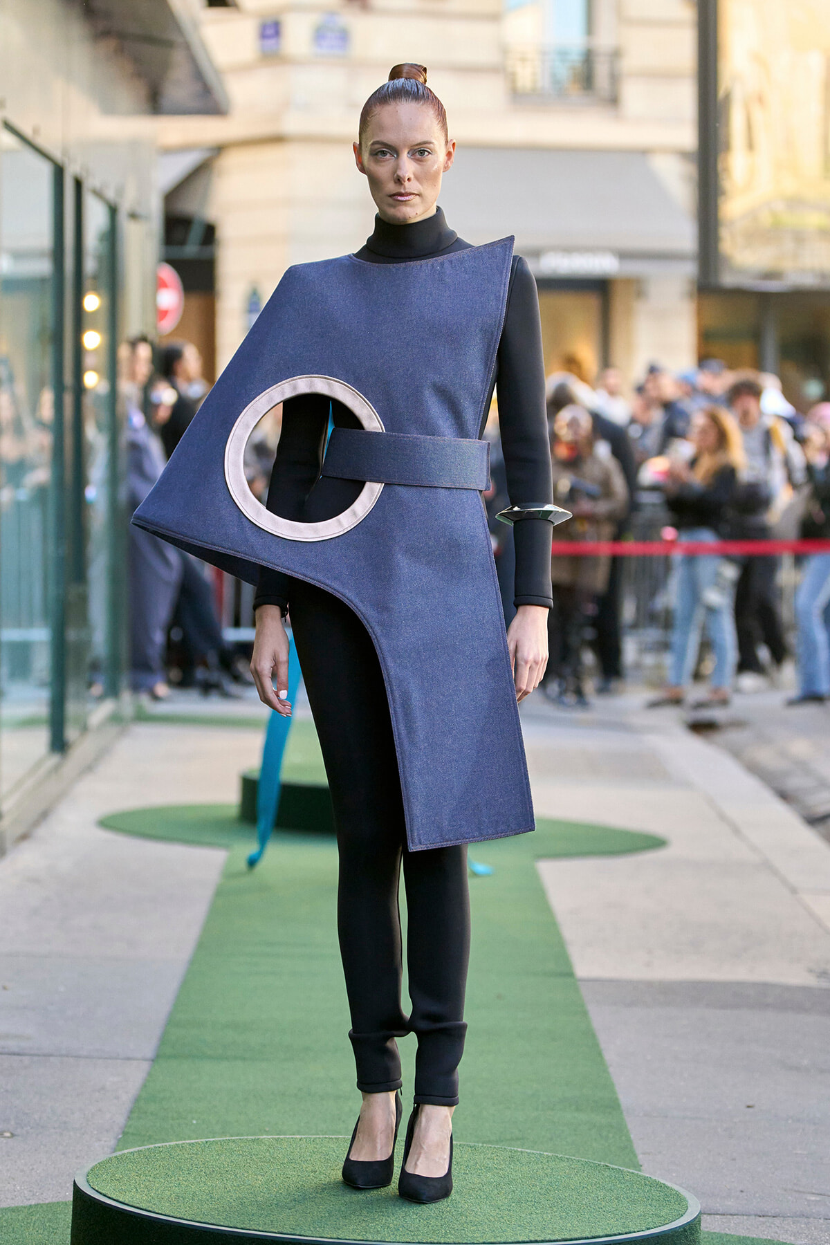 Model wearing an oversized blue geometric dress with a circular cutout, standing on a green runway outdoors.