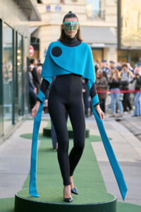 Model in a futuristic turquoise cape over a black bodysuit, wearing a large reflective visor and long turquoise ribbons, standing on a green runway.