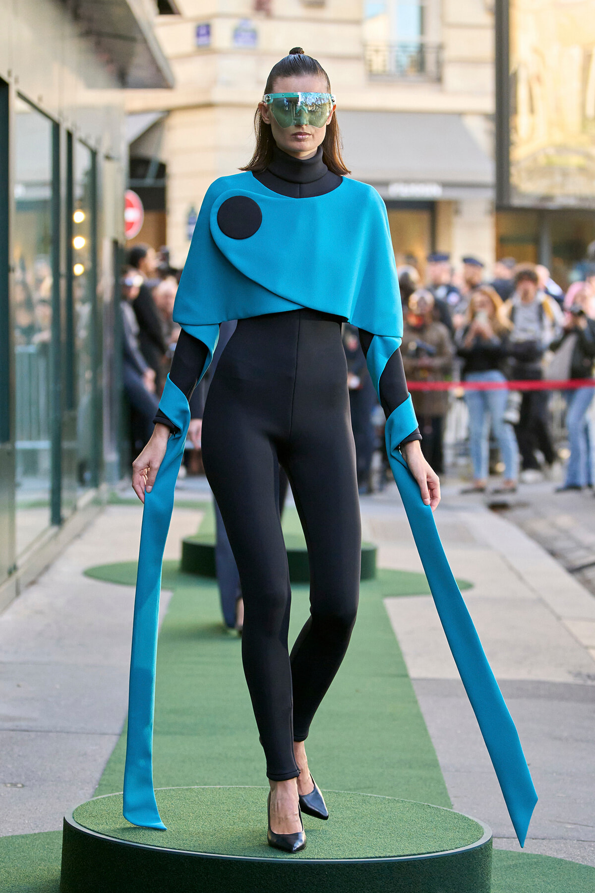 Model in a futuristic turquoise cape over a black bodysuit, wearing a large reflective visor and long turquoise ribbons, standing on a green runway.