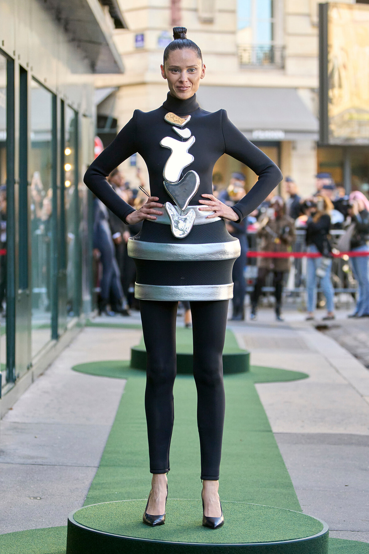 Model wearing a futuristic black dress with stacked metallic shapes on the torso, standing on a green podium at a street fashion show.