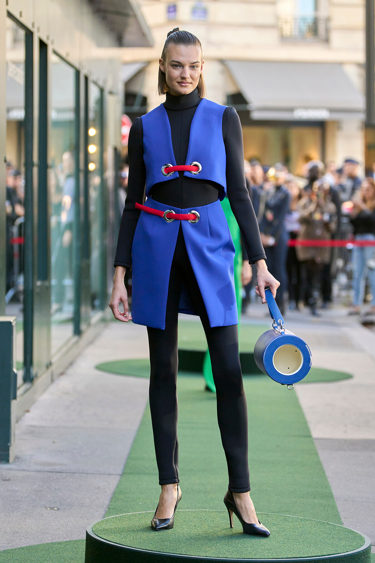 Model wearing a bright blue sleeveless coat with red lacing over a black turtleneck, on a green runway with a blue circular bag in hand.