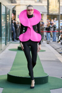 Model walks a green outdoor runway in a neon pink sculptural outfit with oversized circular collar and heart-shaped panels, black bodysuit, pink sunglasses.