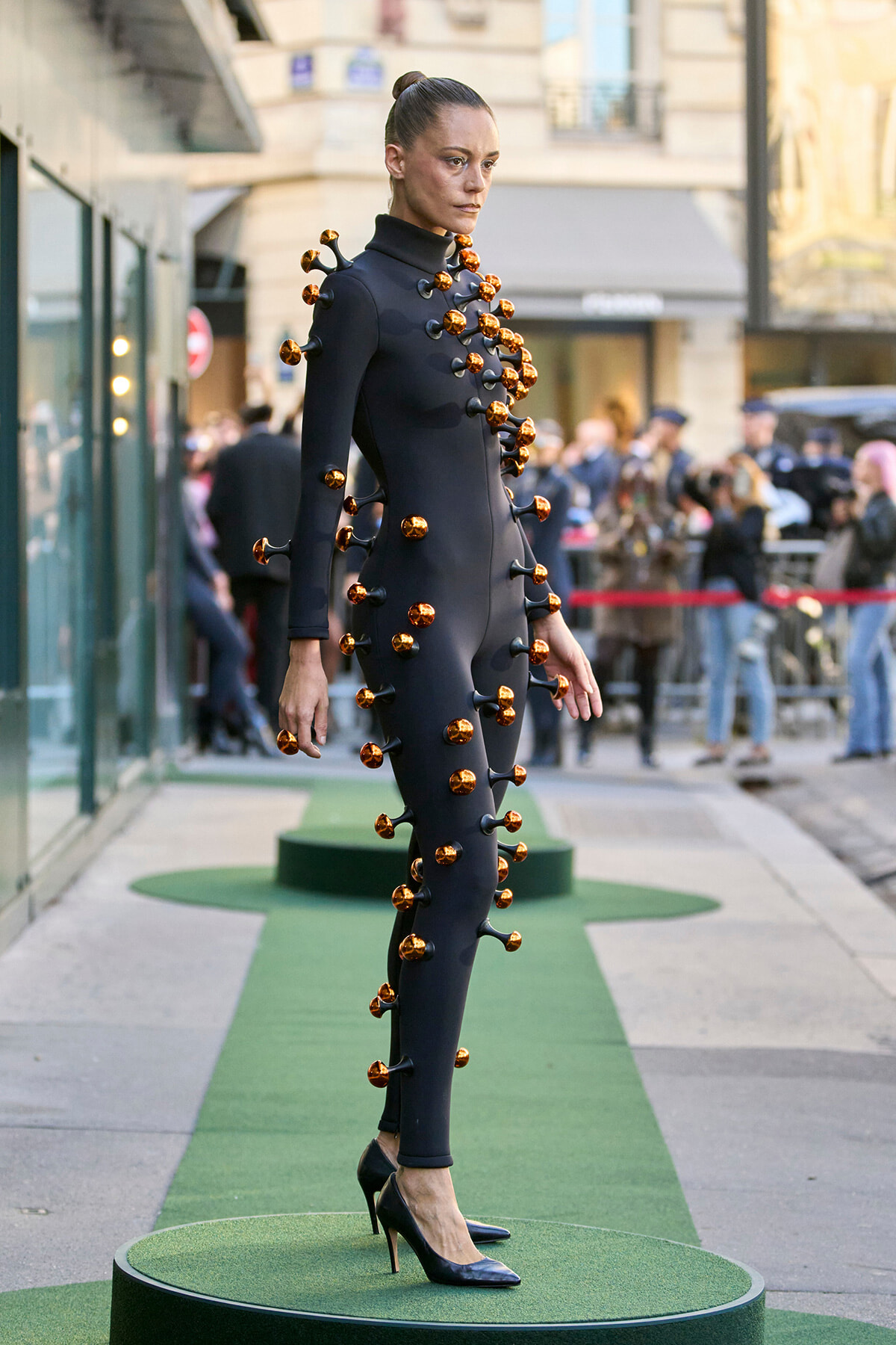Avant-garde model in a black full-body suit with orange knob-like embellishments walking a green runway on a city street.