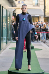 Model on a city sidewalk wearing a blue asymmetrical denim dress over a black bodysuit, with a metallic blue chest ornament.