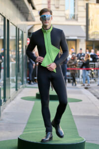 Model wearing a black full-body suit with a neon green angular chest panel, posing on a circular green runway outdoors, sunglasses on.