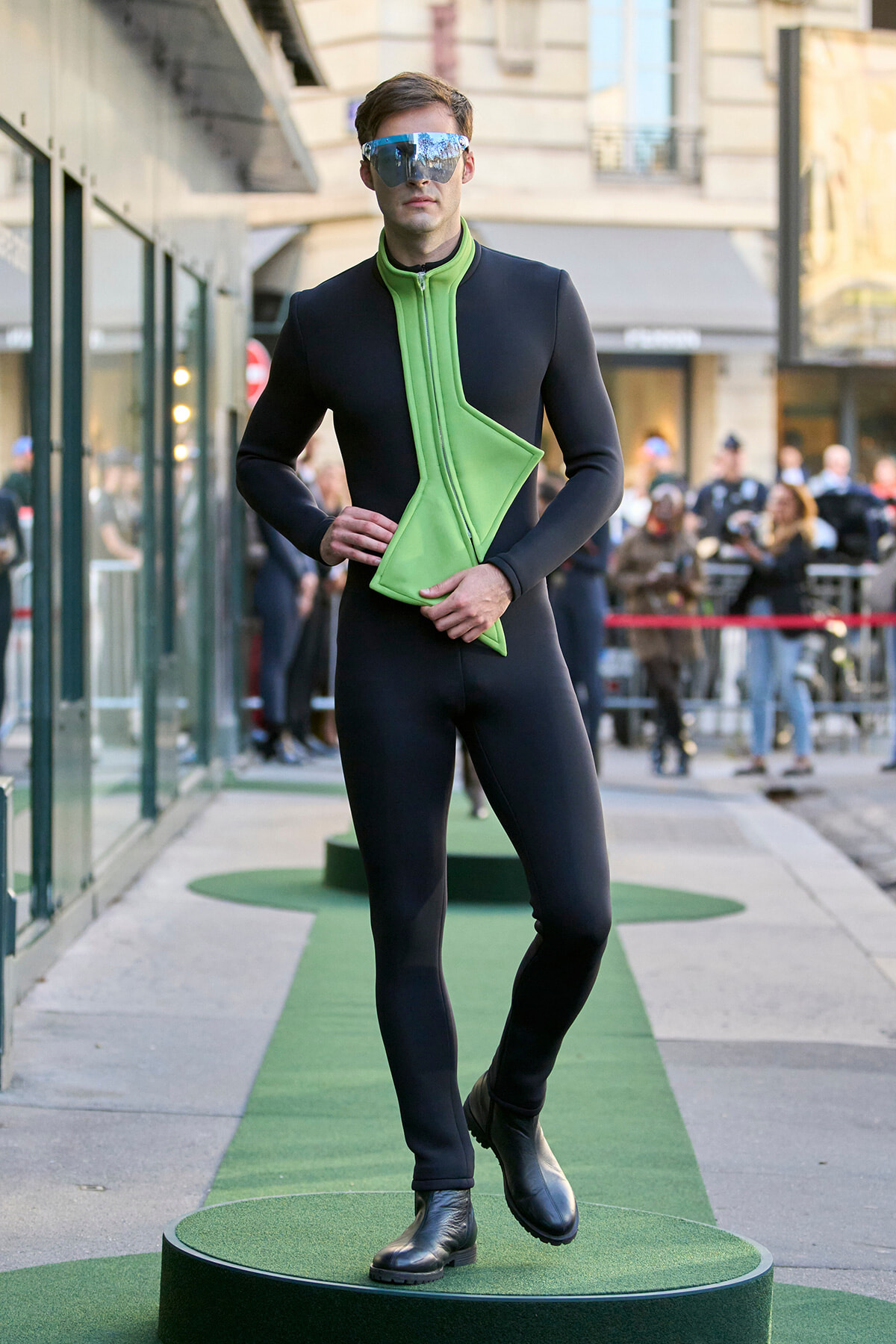 Model wearing a black full-body suit with a neon green angular chest panel, posing on a circular green runway outdoors, sunglasses on.