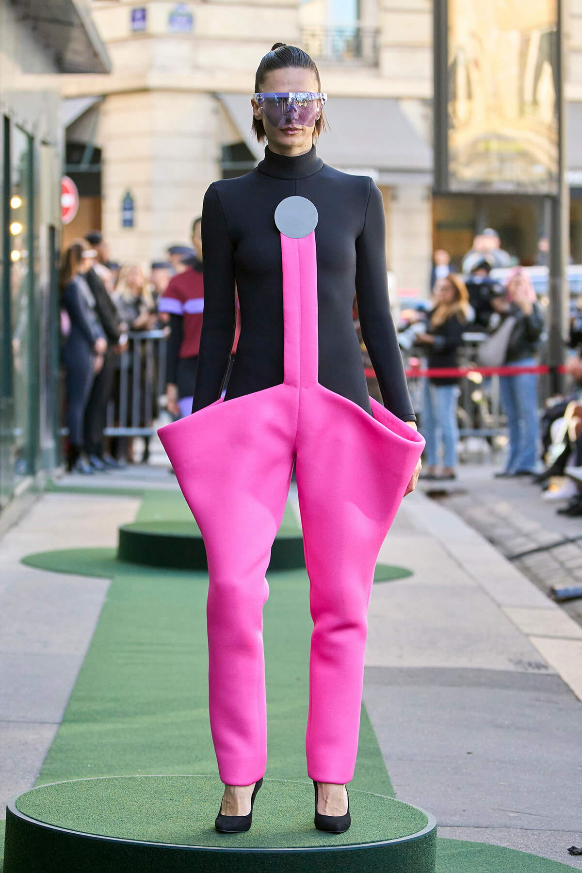 Model wearing a futuristic black bodysuit with bright pink sculpted pants and a gray circular chest piece on a street runway.
