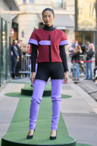 Female model on an outdoor runway wearing a burgundy structured top with lavender bands and matching lilac leggings over a black bodysuit.