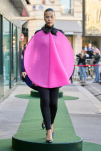 Model wearing a hot pink circular cape over a black outfit walking on a green outdoor runway.