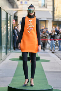 Model poses on a green runway platform in a bright orange sleeveless dress over a black turtleneck and leggings, with a large jeweled cross brooch.