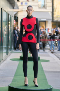 Full-body shot of a model in a bold red and black geometric outfit with circular cutouts, standing on a green circular runway outdoors.