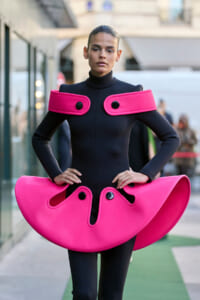 Model wearing a black full-body outfit with large hot-pink sculptural shoulder accents and a matching hip piece, in an urban setting.