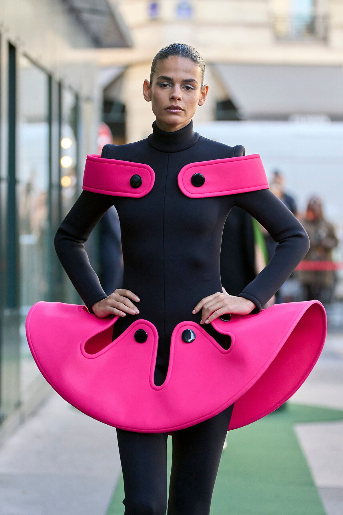 Model wearing a black full-body outfit with large hot-pink sculptural shoulder accents and a matching hip piece, in an urban setting.