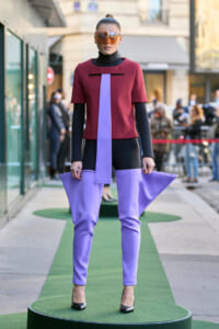 Model posing on a street runway wearing a burgundy top with a lavender sash and matching lavender pants, plus oversized orange sunglasses on a green platform.