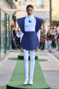 Model wearing a futuristic blue and lavender outfit with oversized shoulders, standing on a circular green runway in an urban street setting.