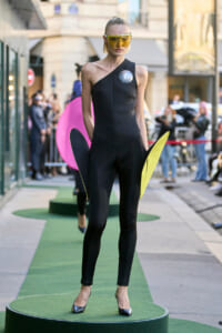 Model on a green runway wearing a one-shoulder black jumpsuit, large yellow visor sunglasses, and holding neon wing props (yellow and pink).