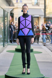 Model wearing a bold purple geometric top with blue panels over a black outfit, standing on a green runway outdoors.