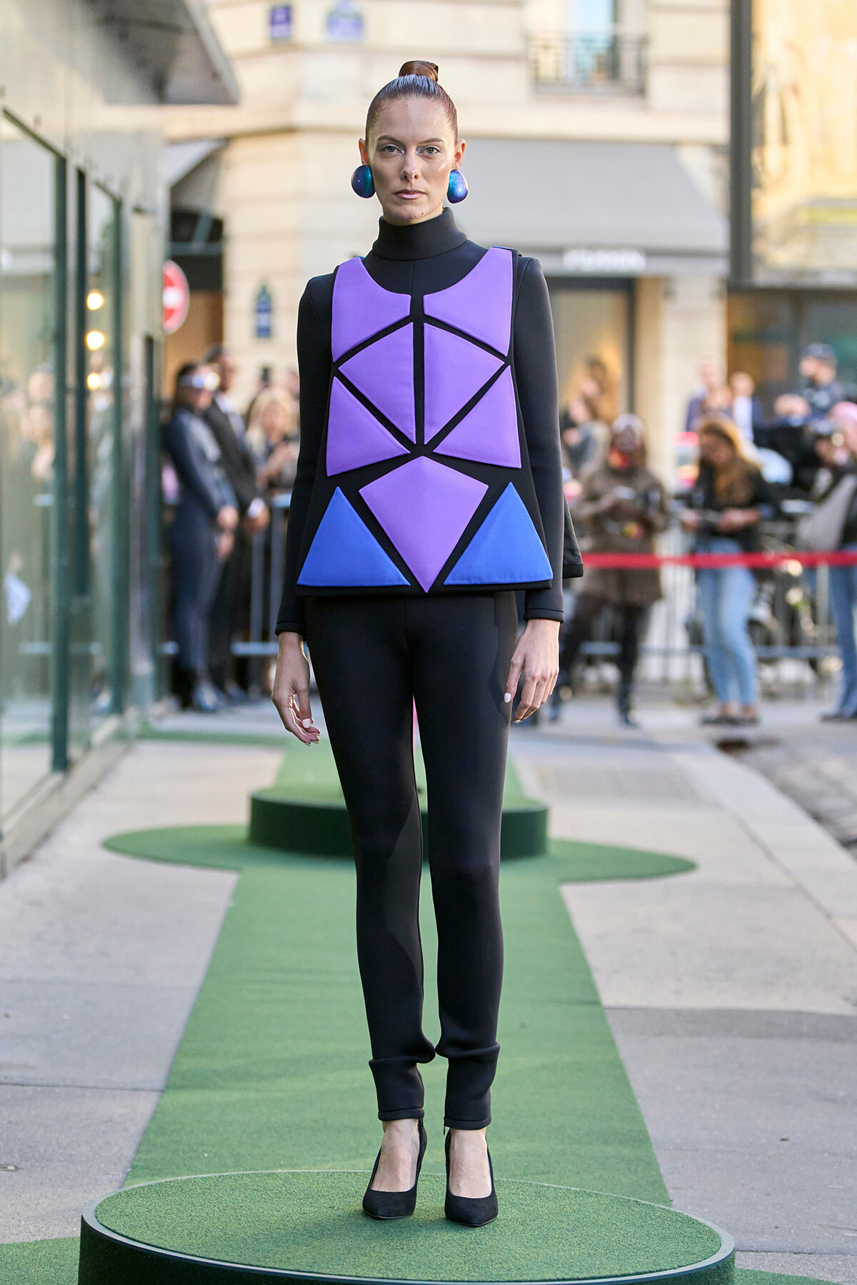 Model wearing a bold purple geometric top with blue panels over a black outfit, standing on a green runway outdoors.