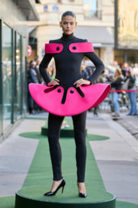 Model wearing a black bodysuit with large pink shoulder and hip accessories during a street fashion show on a green runway.