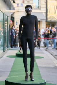 Model in a black full-body catsuit and high heels stands on a green runway outdoors, with a crowd watching in the background.