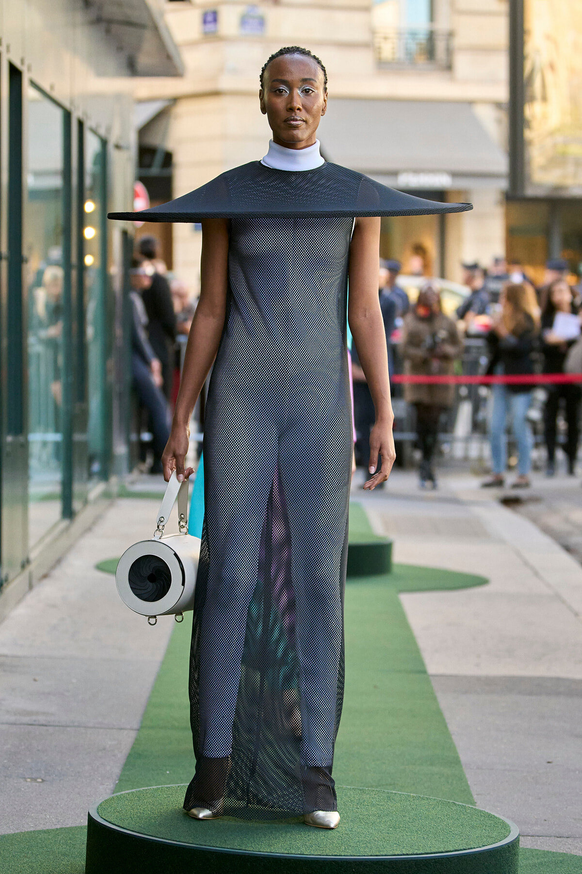 Model in a futuristic black perforated jumpsuit with a wide, flat circular hat on a green runway outdoors, holding a round white bag.