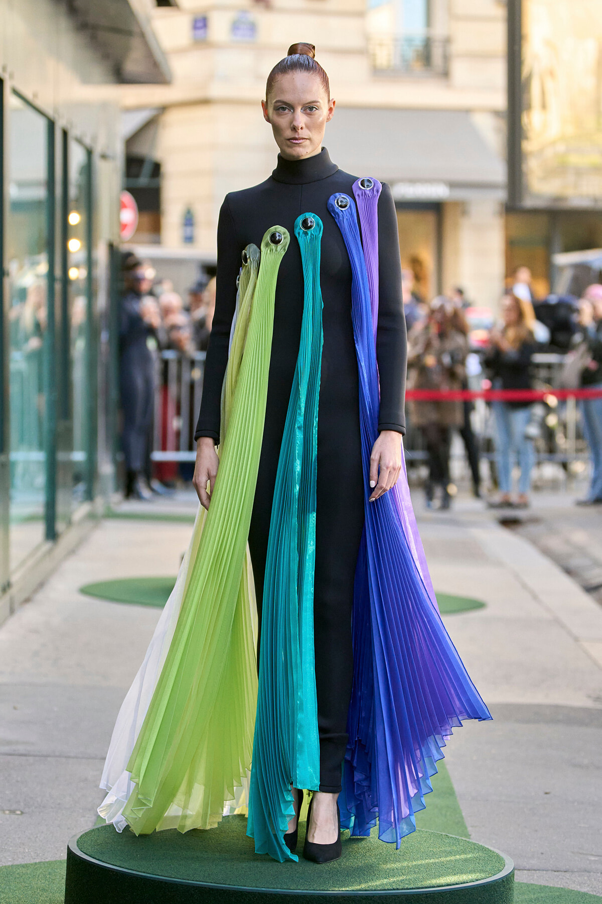 Model in a black turtleneck gown with long lime, teal, turquoise, and purple pleated ribbons cascading from shoulders on a runway platform.