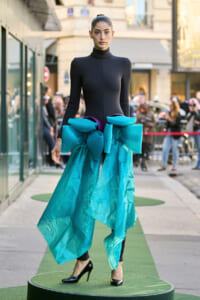 Model on a street runway wearing a black turtleneck bodysuit with a large turquoise bow and flowing teal fabric at the hips, paired with black heels.
