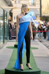 Model on a city street wearing a futuristic blue and black gown with a large foam sculpture draped across the chest and yellow visor-style sunglasses.