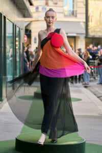 Model wearing a bold color-block gown with sheer black overlay, standing on a green circular platform at an outdoor street fashion show, holding the fabric to create movement, with a red circular accessory at the chest.