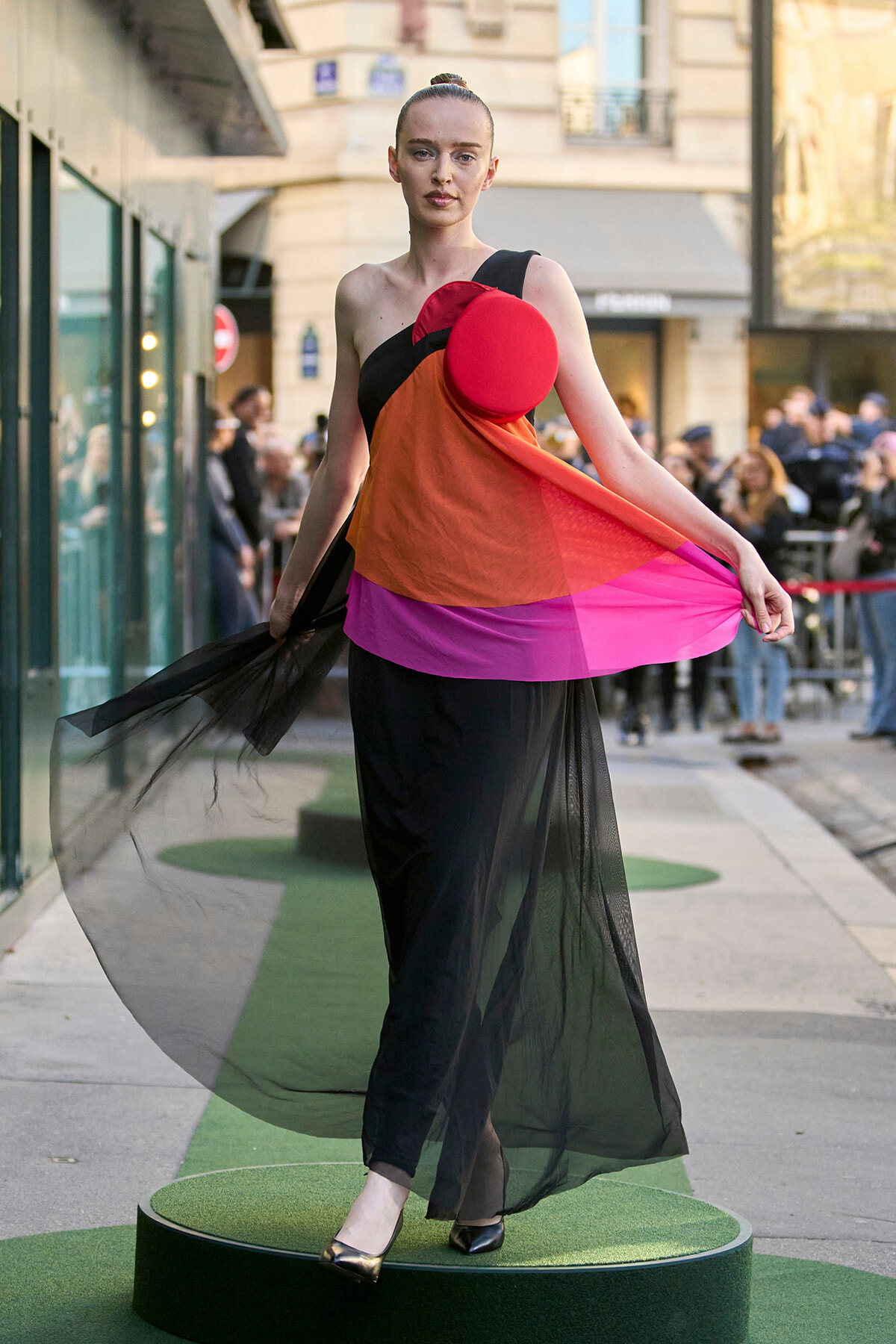 Model wearing a bold color-block gown with sheer black overlay, standing on a green circular platform at an outdoor street fashion show, holding the fabric to create movement, with a red circular accessory at the chest.
