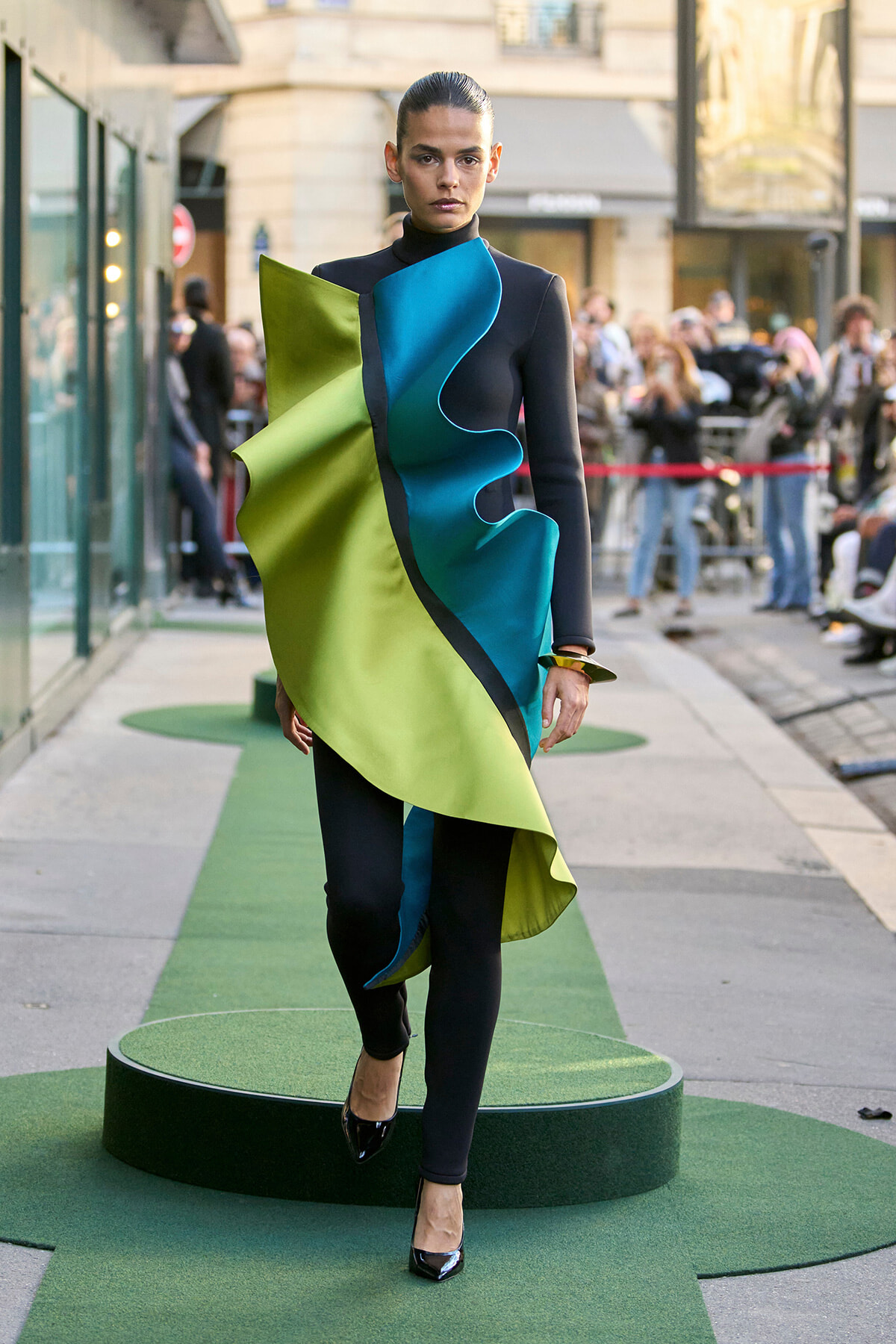 Model walks a city street runway in a avant-garde lime green and teal sculptural dress over black bodysuit, with onlookers in the background.