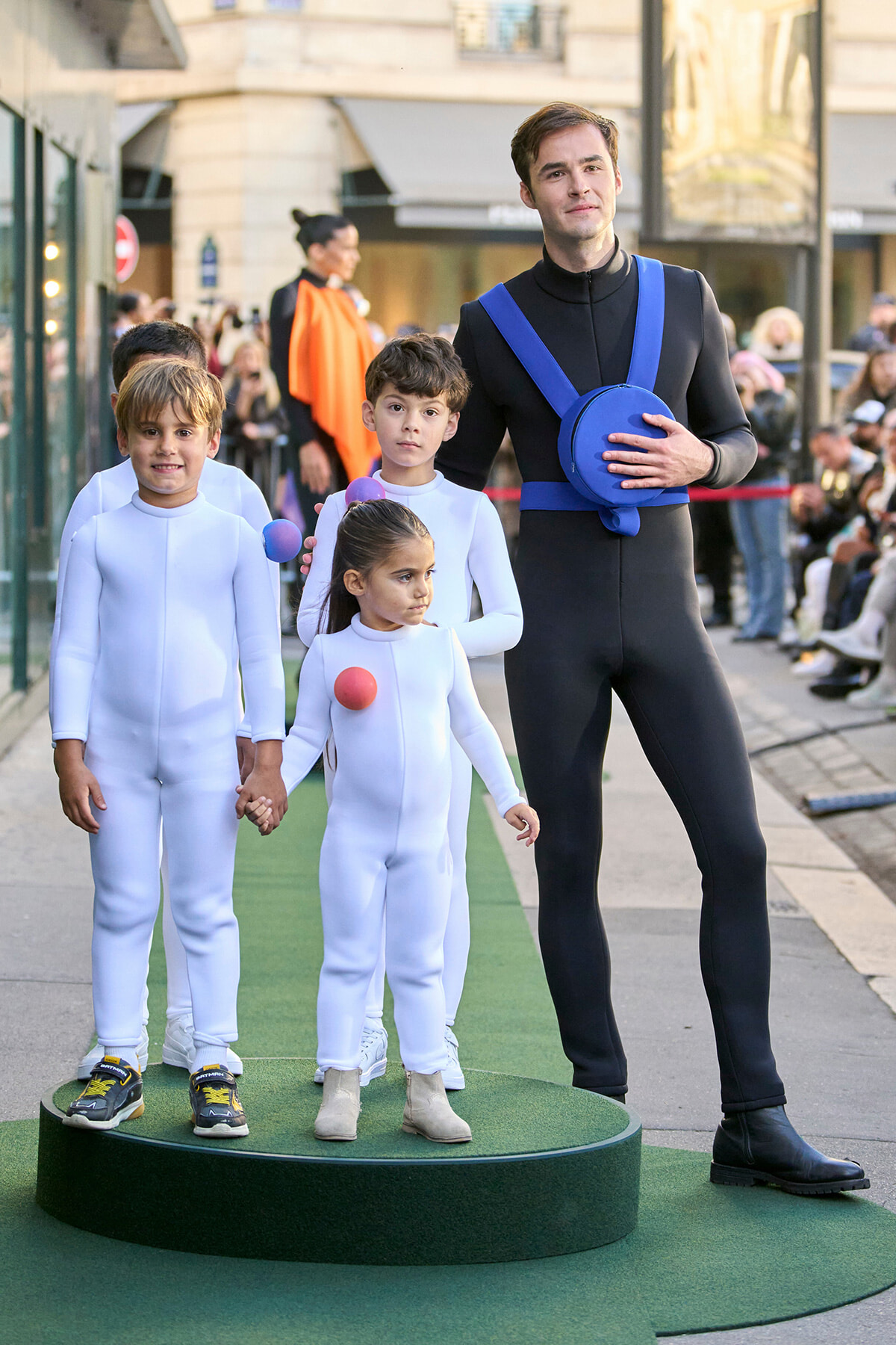 Man in a black outfit with a blue harness holds a blue hat while standing on a green podium with three children in white suits at an outdoor event.