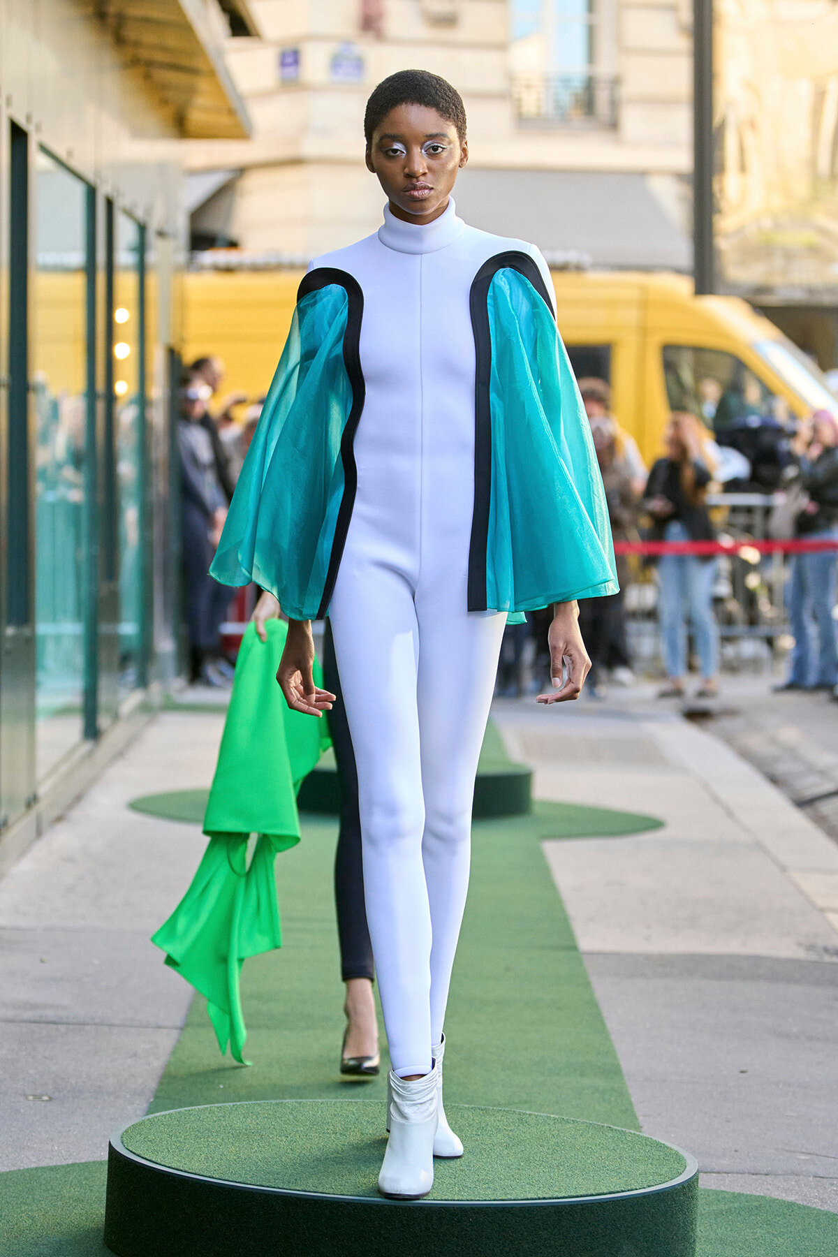 Model walking a green catwalk on a street, wearing a white jumpsuit with teal, cape-like sleeves trimmed in black.