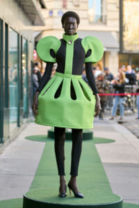 Female model wearing a lime green and black sculptural dress with oversized shoulder shapes, on a circular green platform at a street fashion show.