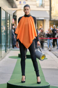 Model wearing an orange draped cape over a black bodysuit, standing on a green circular platform at a street fashion show.