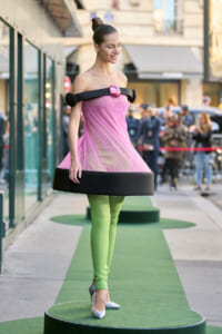 Model on a circular green platform wearing a pink off-shoulder pleated dress with a black band, lime tights, and silver heels.