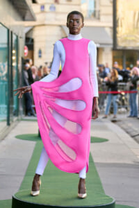 Model in a bright pink sculptural dress with cutout panels over a white bodysuit, posing on a green outdoor runway with spectators in the background.