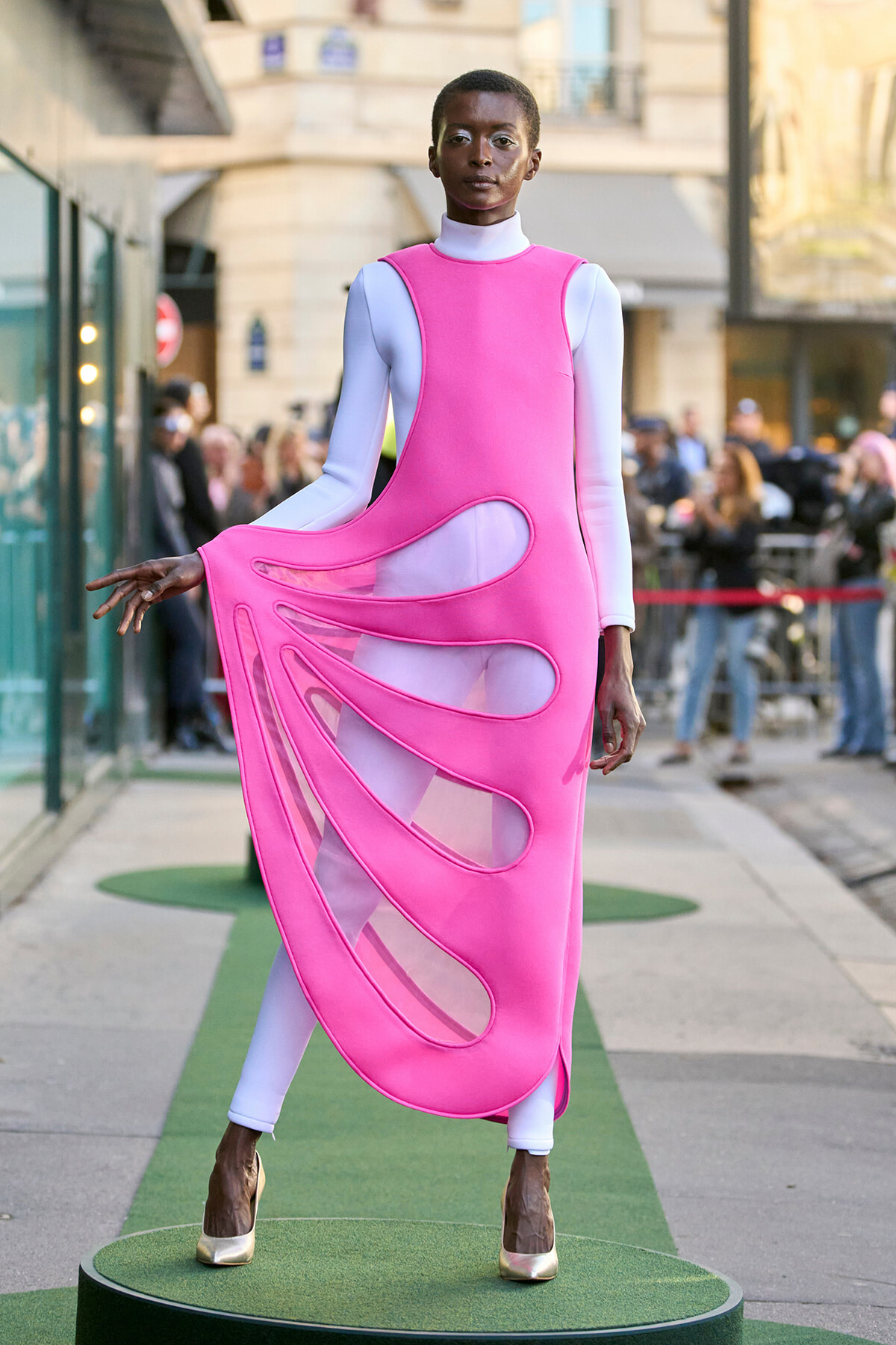 Model in a bright pink sculptural dress with cutout panels over a white bodysuit, posing on a green outdoor runway with spectators in the background.