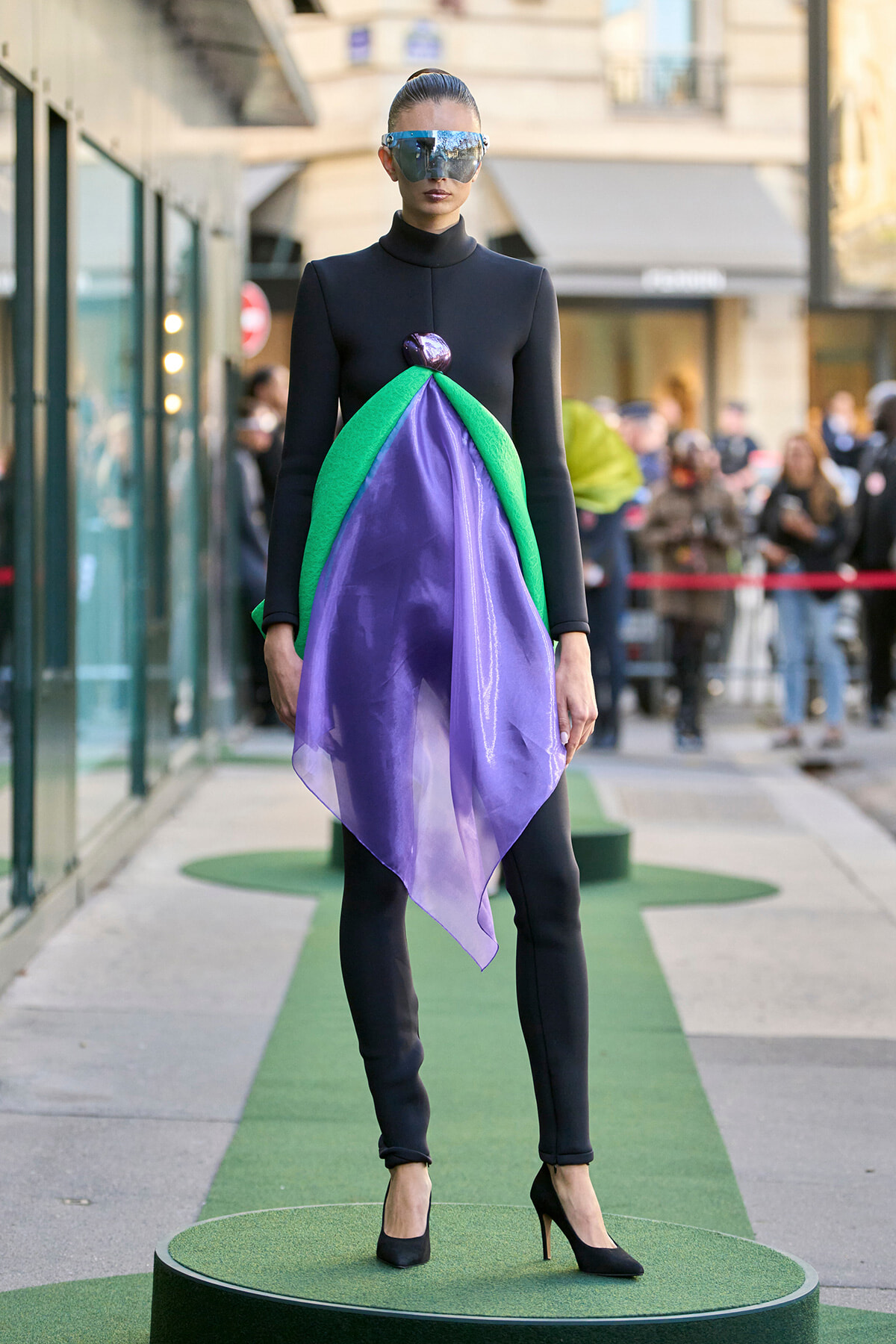 Model in a black long-sleeve bodysuit wearing a purple, green draped avant-garde dress panel on a green runway outdoors, with reflective futuristic sunglasses.