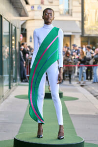 Model on an outdoor runway wearing a white bodysuit with a bold green diagonal sash accented by pink and blue stripes; spectators in the background.