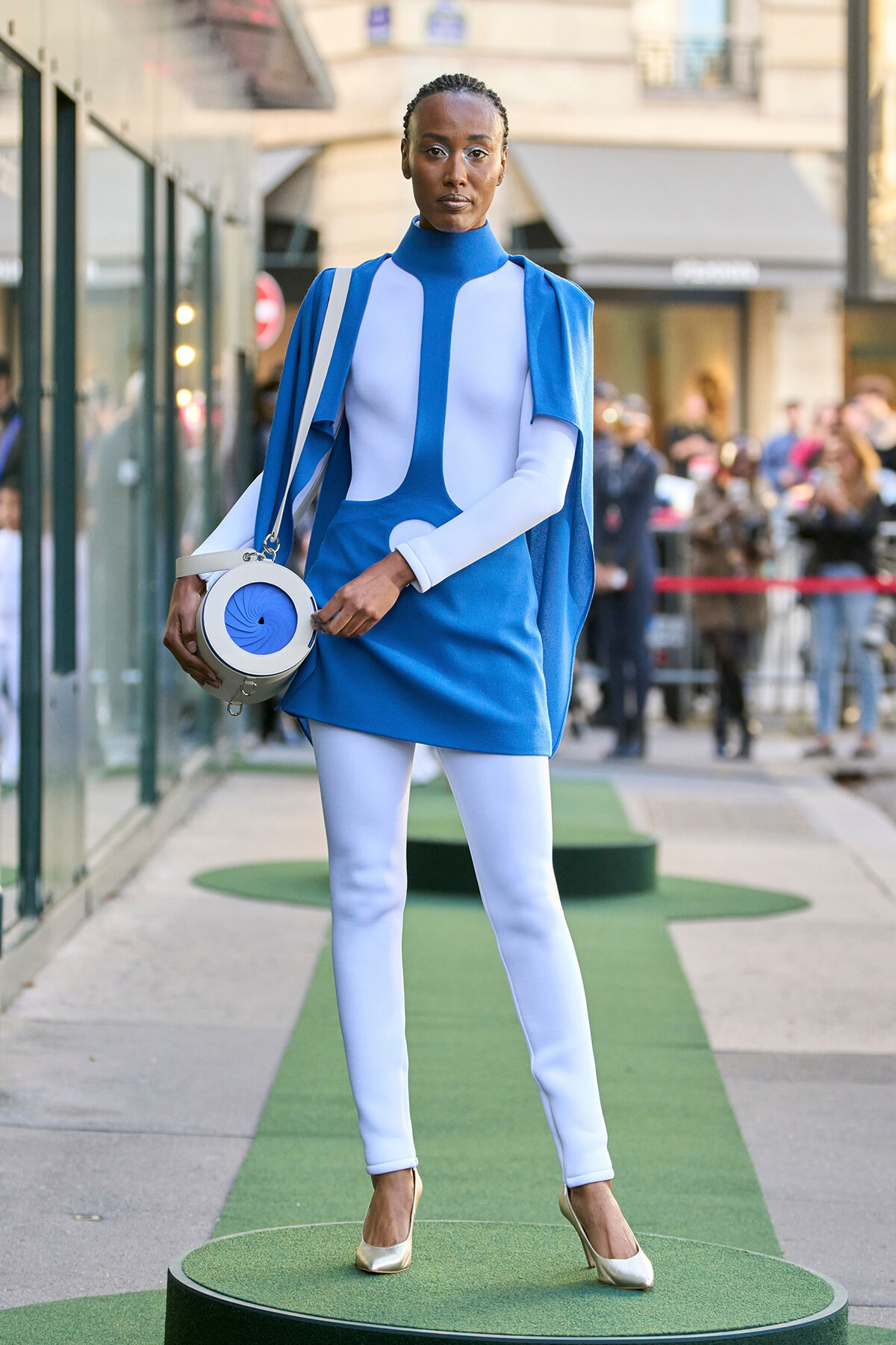 Model posing on a street runway in a blue and white structured futuristic dress with a cape, white leggings, gold heels, and a round white-and-blue handbag, standing on a green circular platform while spectators watch in the background.