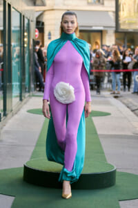 Model walks a lavender bodysuit with teal cape and oversized white flower at the hip on a green runway, with a street crowd in the background.