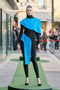 Model on an outdoor runway wearing a black bodysuit with bright blue geometric cape and panel, standing on a green platform with onlookers in the background