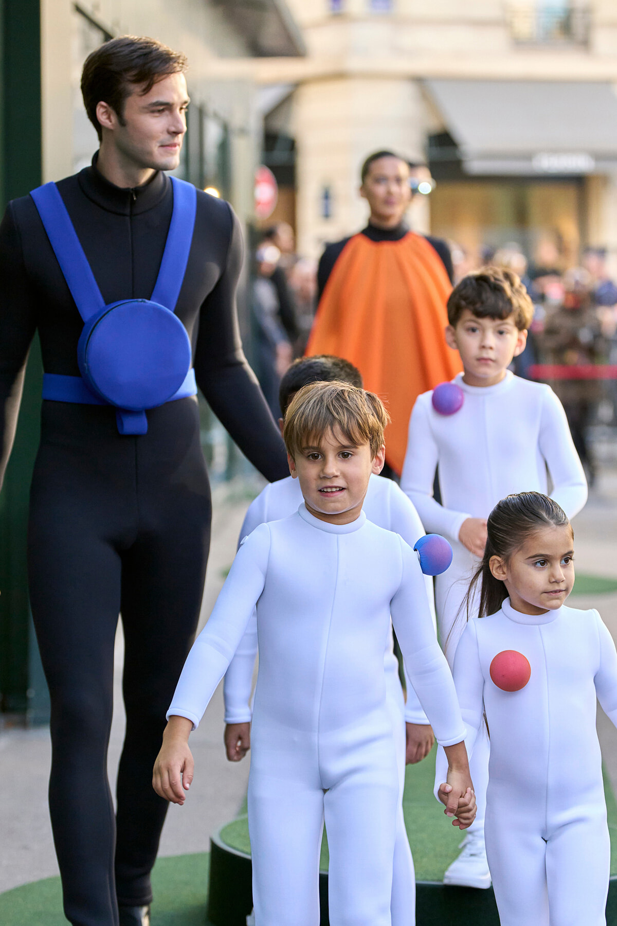 A man in a black suit with a blue harness walks with several children in white bodysuits during an outdoor event, holding hands.